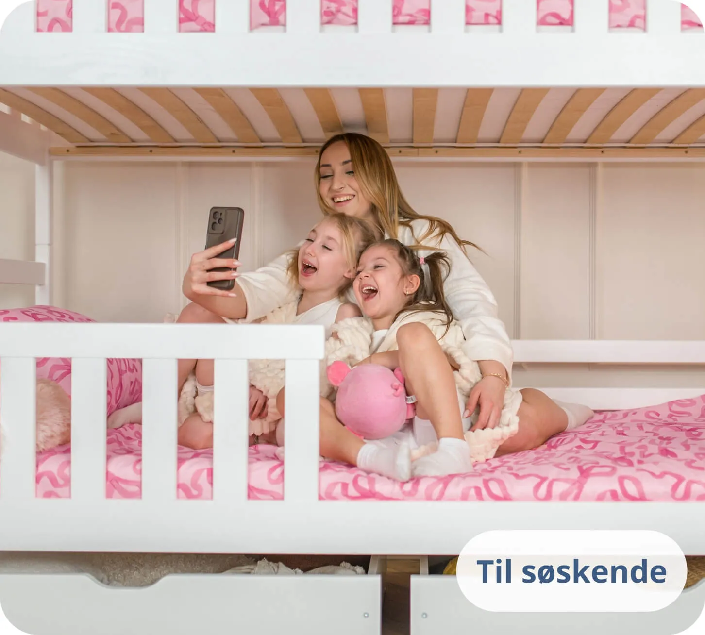 To piger og en kvinde griner ad en telefon på en hvid køjeseng.